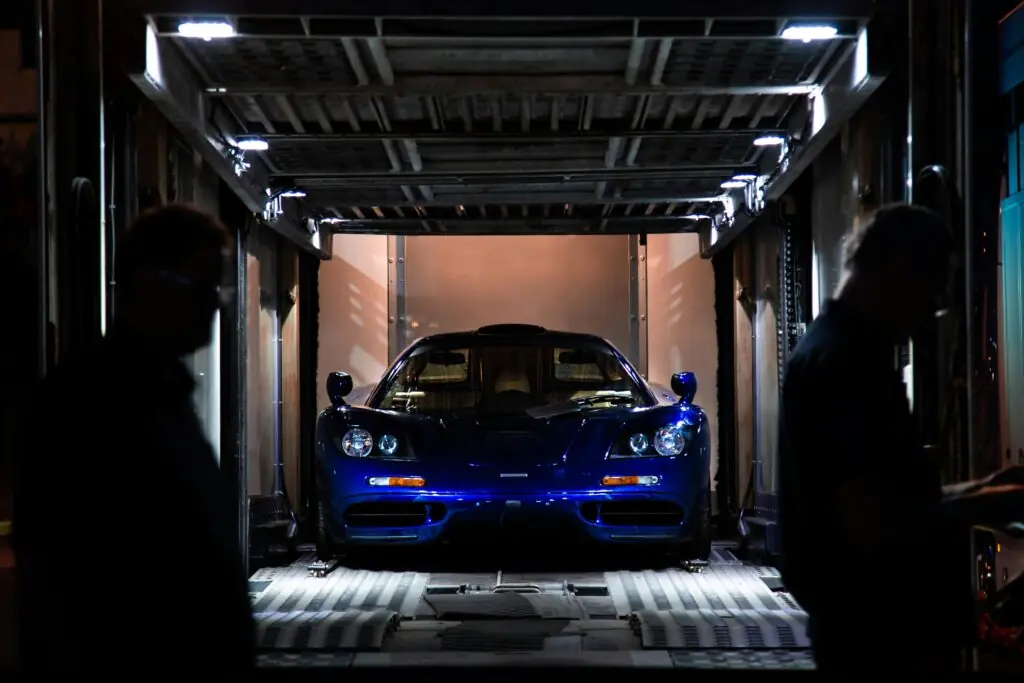 A sleek blue sports car is partially illuminated inside a shipping container, with silhouettes of two people in the foreground, hinting at its valuable transport.