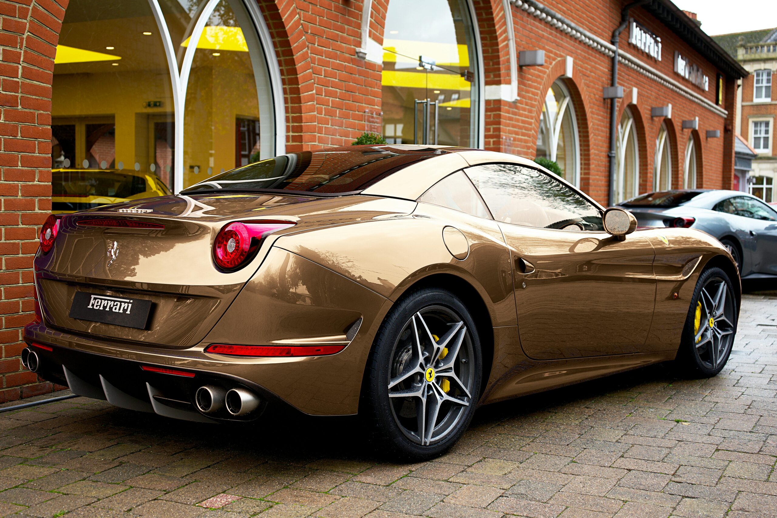 A sleek brown Ferrari convertible parked on brick pavement outside a dealership, showcasing its elegant curves and distinctive design.